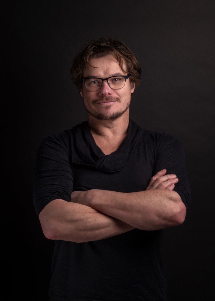Portret van Frank de Bruijn, met bril en armen over elkaar, tegen een donkere achtergrond.