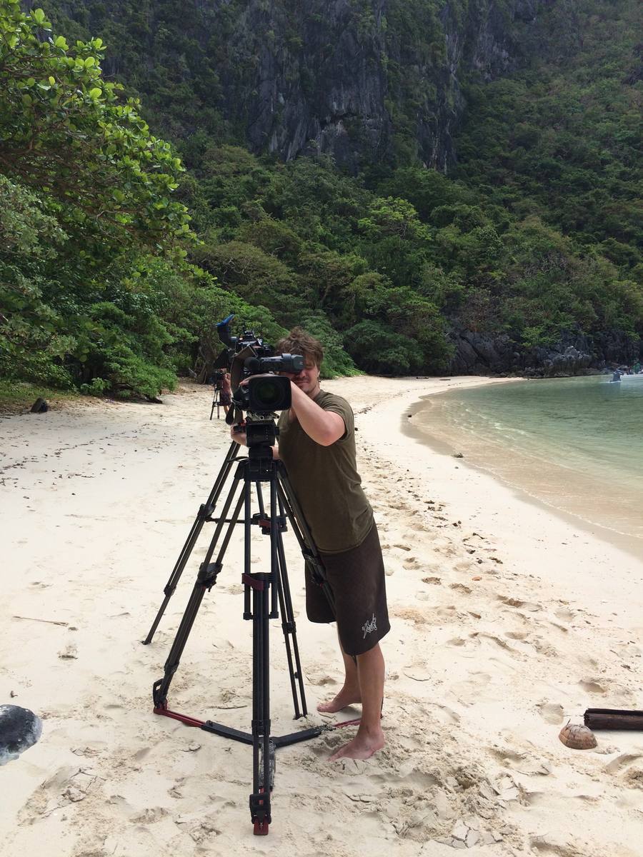 Frank filmt met een schoudercamera op statief op een tropisch strand, met begroeide kliffen en een baai op de achtergrond.